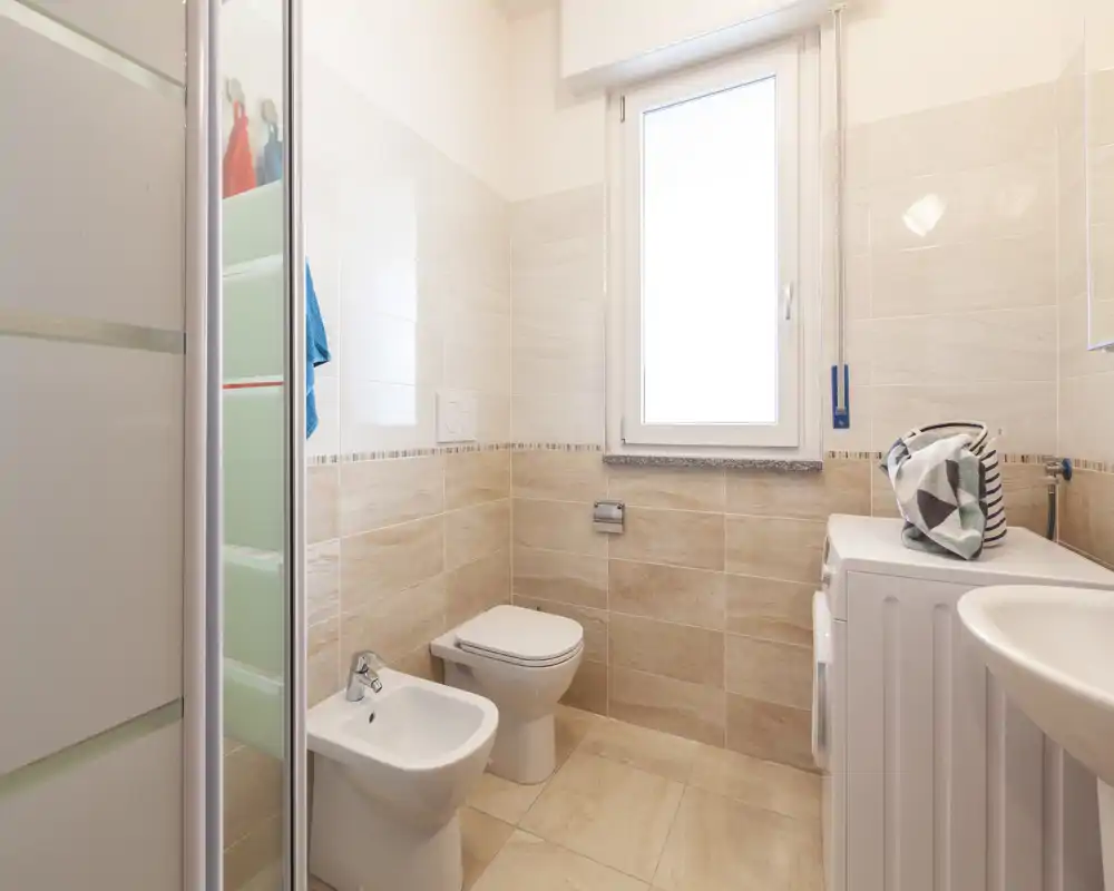 Bagno moderno e luminoso con piastrelle, bidet, WC, box doccia e lavabo con lavatrice — pulito e ben illuminato.