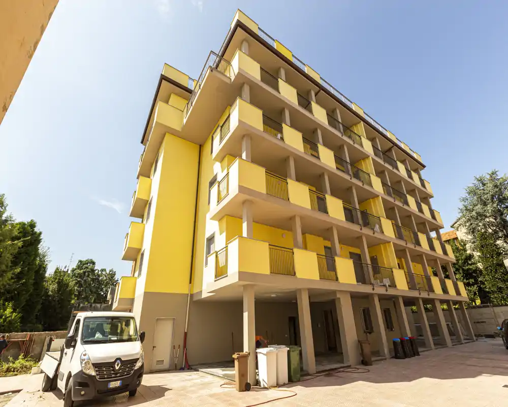 Vista esterna luminosa di un edificio residenziale recentemente tinteggiato di giallo con più balconi e un cortile pavimentato, ripreso in pieno giorno.