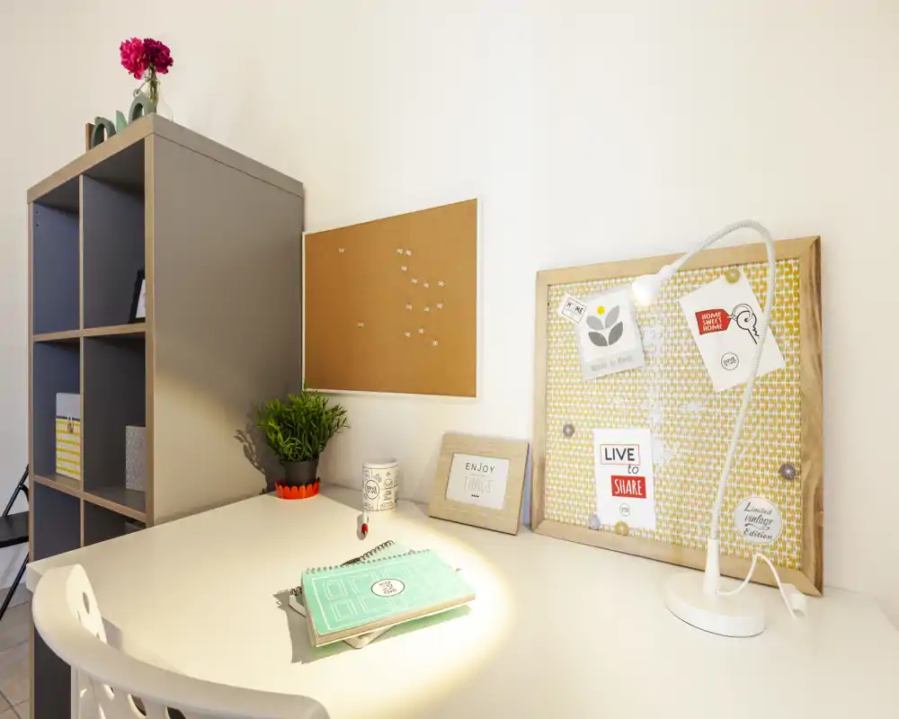 Spazio di lavoro domestico luminoso e ordinato con scrivania bianca, pannelli in sughero decorativi, lampada da tavolo e pianta — perfetto per studio o smart working.
