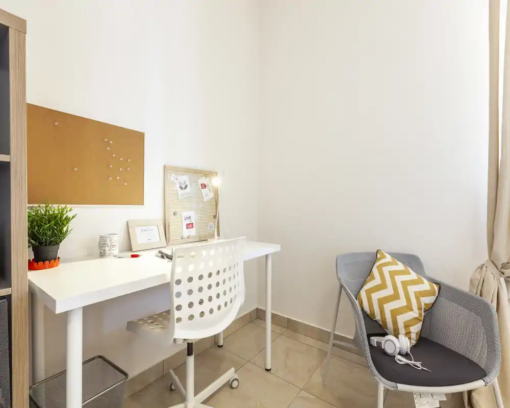 Angolo studio compatto e luminoso con scrivania bianca, sedia girevole forata, bacheca in sughero e una poltrona d'accento con cuscino a zigzag — perfetto per lavorare o studiare.