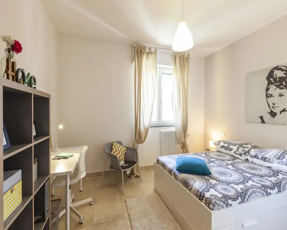 Camera da letto luminosa e moderna con letto matrimoniale, biancheria decorata, lampada da comodino e un angolo studio con scrivania e sedia. La finestra con tende leggere fa entrare luce naturale e la palette neutra crea un'atmosfera tranquilla.
