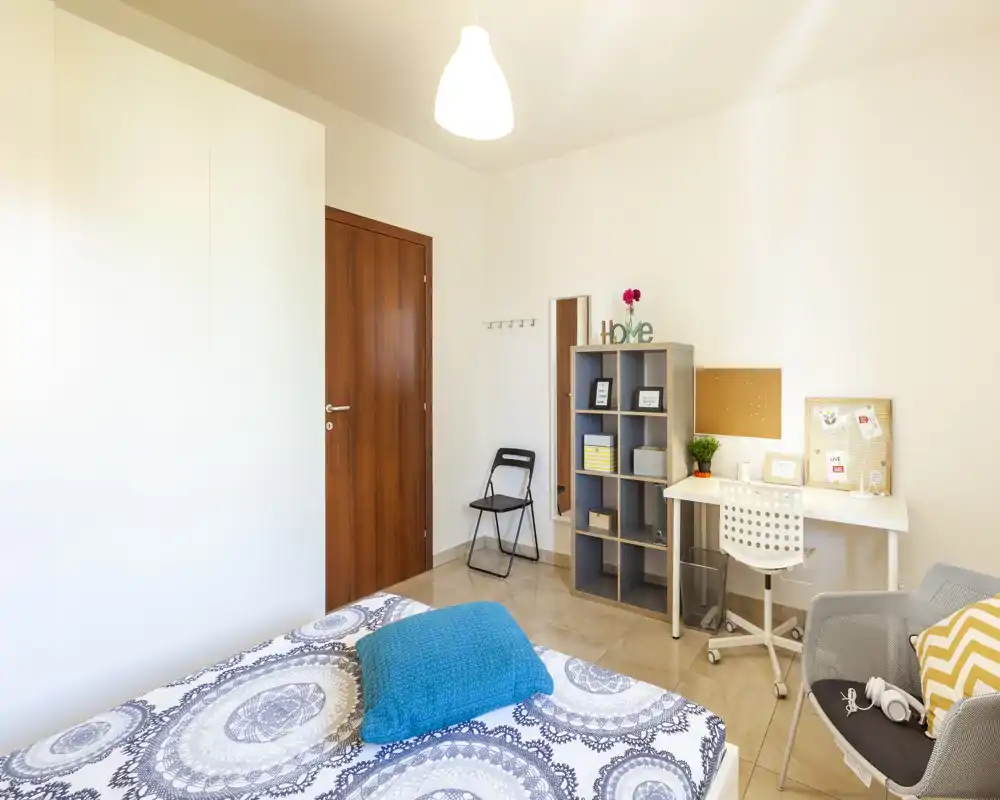 Camera da letto luminosa e ben organizzata con copriletto decorato, cuscino turchese, scrivania e scaffale: ambiente accogliente e funzionale per riposo e lavoro.