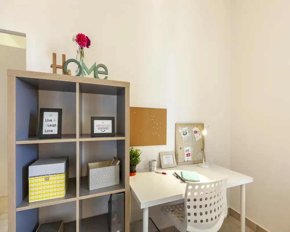 Angolo di lavoro domestico luminoso e ordinato con scrivania bianca, sedia e scaffale organizzato: cornici decorative, pianta e bacheca rendono lo spazio accogliente e funzionale.