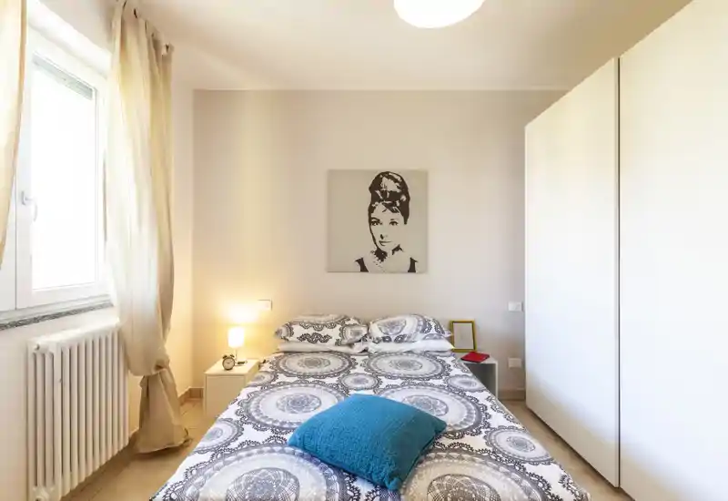 Camera da letto luminosa e ordinata con letto matrimoniale al centro sotto un quadro, luce naturale morbida dalla finestra e grande armadio per riporre.