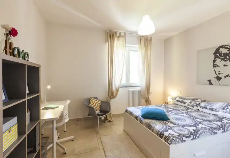 Dormitorio luminoso y moderno con cama doble, ropa de cama estampada, lámpara de mesita y un espacio de trabajo con escritorio y silla. La ventana con visillos aporta luz natural y la paleta neutra crea un ambiente relajado.