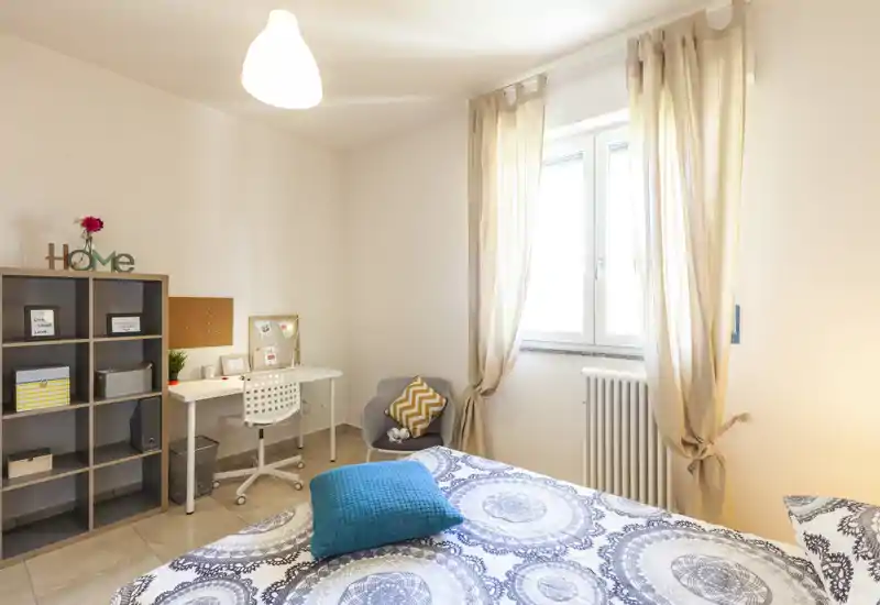 Camera da letto luminosa e ben arredata con letto matrimoniale dal motivo, cuscini confortevoli, angolo studio e scaffalatura—ideale per un soggiorno singolo o di coppia con luce naturale.