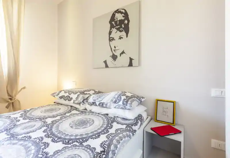 Camera da letto luminosa e di stile con un letto matrimoniale con motivo, tessili morbidi e un ritratto incorniciato sopra la testiera. La luce naturale entra da sinistra, creando uno spazio accogliente e ben curato.