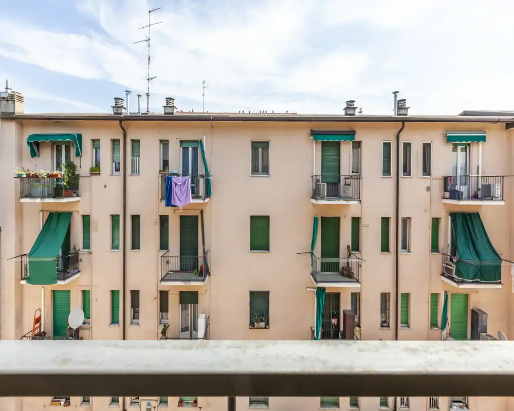 Vista dal balcone verso la facciata di un edificio residenziale con piccoli balconi e persiane verdi; cielo luminoso e dettagli architettonici rendono la scena un valido esterno urbano.