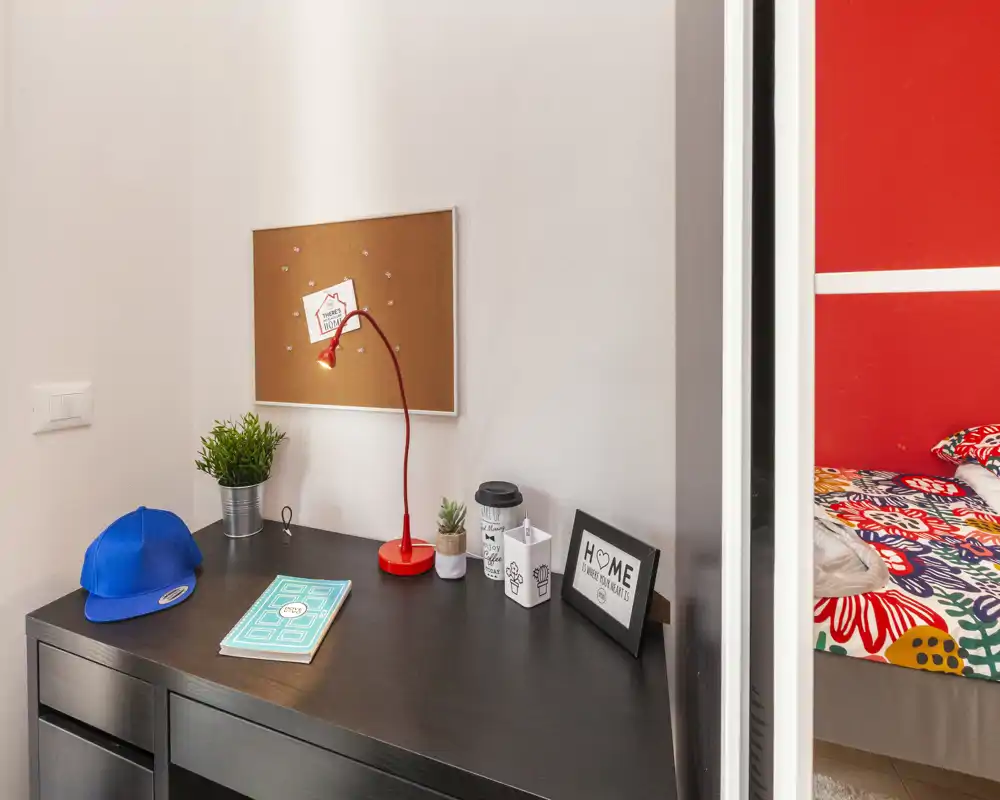 Veduta della camera da letto focalizzata sulla scrivania con parziale vista del letto colorato a destra. La scrivania ha una lampada rossa, una pianta, un quaderno e accessori in un ambiente luminoso e ordinato.