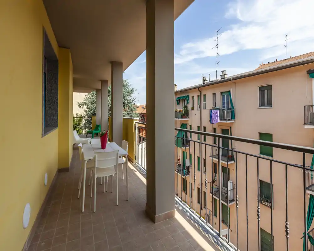 Balcone soleggiato arredato con tavolo e sedie, pavimento in piastrelle e ringhiera che si affaccia sugli edifici vicini — ideale per pranzi all'aperto e relax.