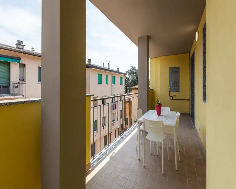 Balcone coperto e soleggiato con tavolo e sedie, affacciato sugli edifici vicini — perfetto per pranzi all'aperto e relax.