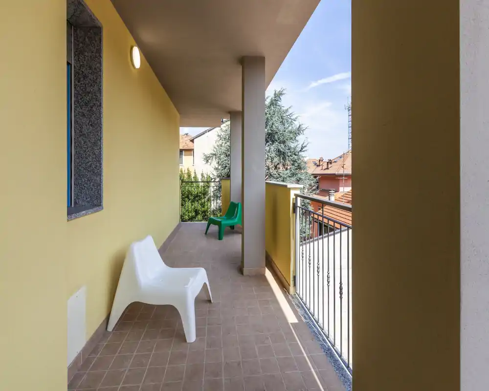 Balcone coperto soleggiato con pavimento piastrellato, sedie in plastica semplici e ringhiera in metallo che si affaccia sui tetti vicini—ideale per rilassarsi all'aperto.