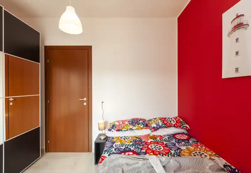 Camera da letto luminosa e colorata con letto matrimoniale davanti a una parete rossa, biancheria a fantasia, comodino con lampada e armadio — ambiente accogliente e vivace perfetto per annunci.