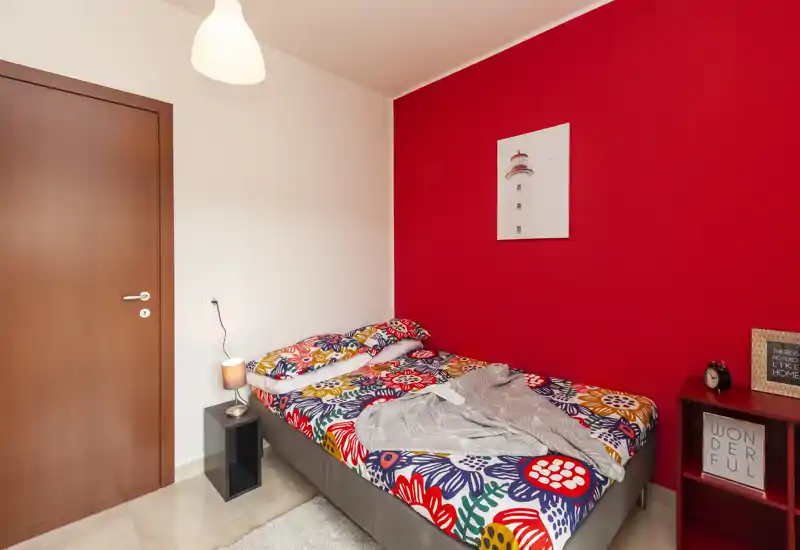 Camera da letto compatta e luminosa con un letto matrimoniale colorato davanti a una parete rossa vivace, lampada da comodino e mensola semplice — ideale per un soggiorno accogliente.