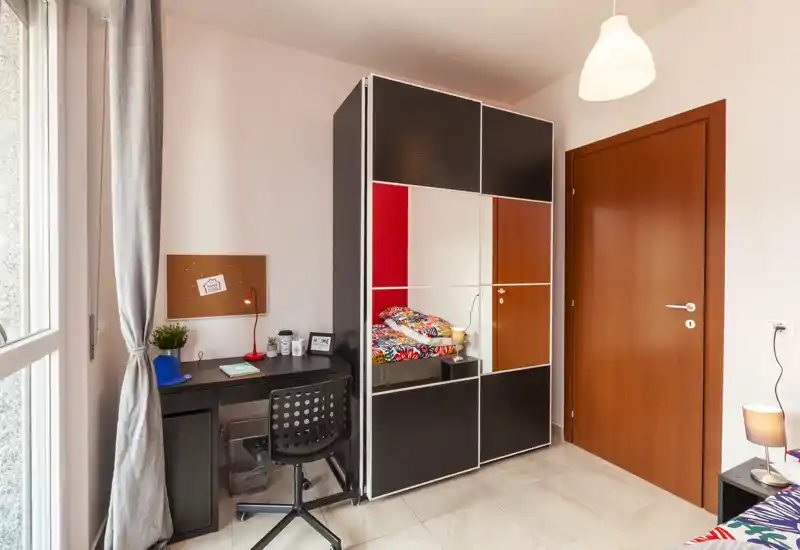 Angolo di camera luminoso e moderno con armadio con specchio, scrivania compatta e letto colorato parzialmente visibile; buona luce naturale e stile ordinato, adatto ad affitti.