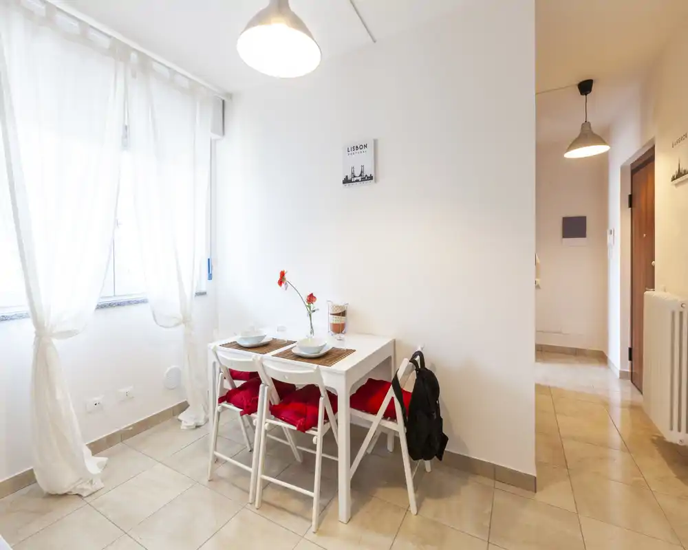 Angolo pranzo luminoso e compatto con un piccolo tavolo bianco apparecchiato e sedie pieghevoli con cuscini rossi, luce naturale soffusa dalla finestra e arredo minimale.