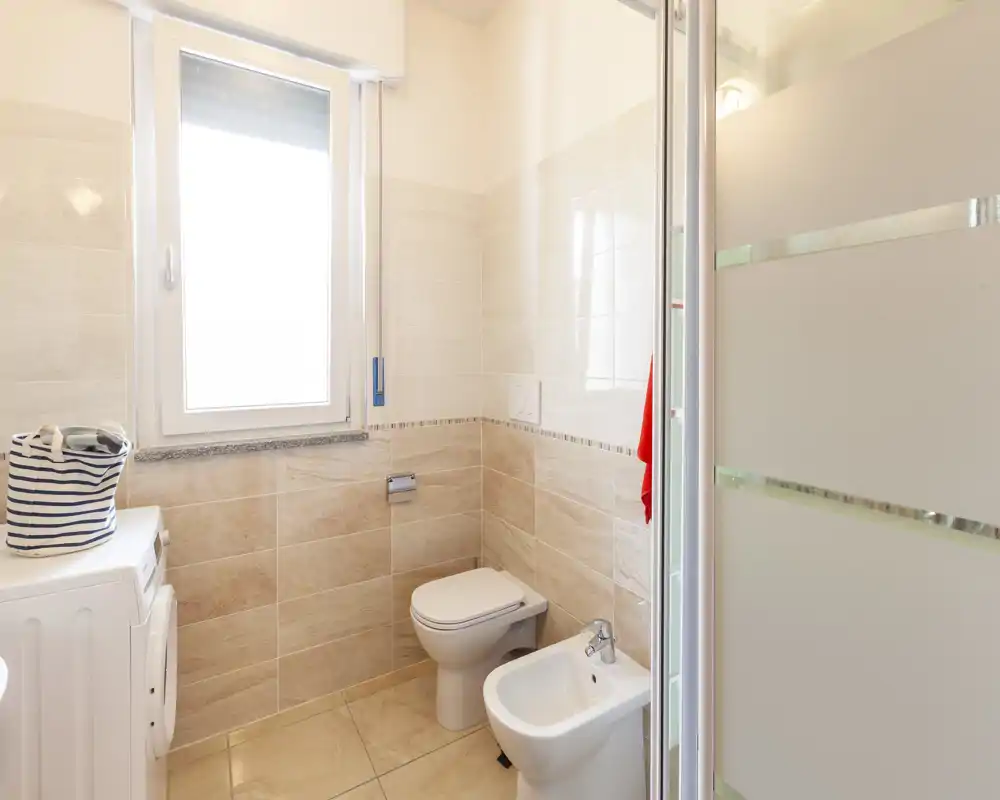 Bagno moderno e luminoso con wc a parete, bidet e box doccia in vetro. La luce naturale dalla finestra e le piastrelle beige chiaro donano un aspetto pulito e fresco.