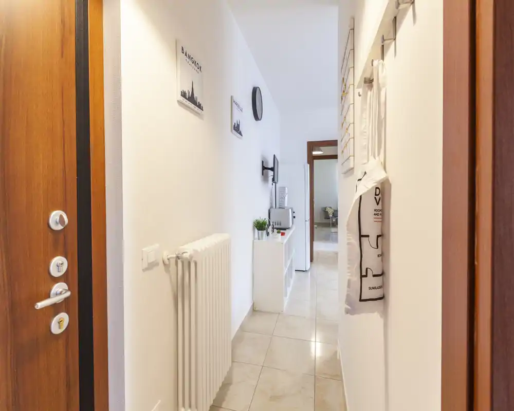 Ingresso luminoso e ordinato con appendiabiti a parete, termosifone e un piccolo mobile console lungo il corridoio — pavimento in piastrelle e arredi neutri.