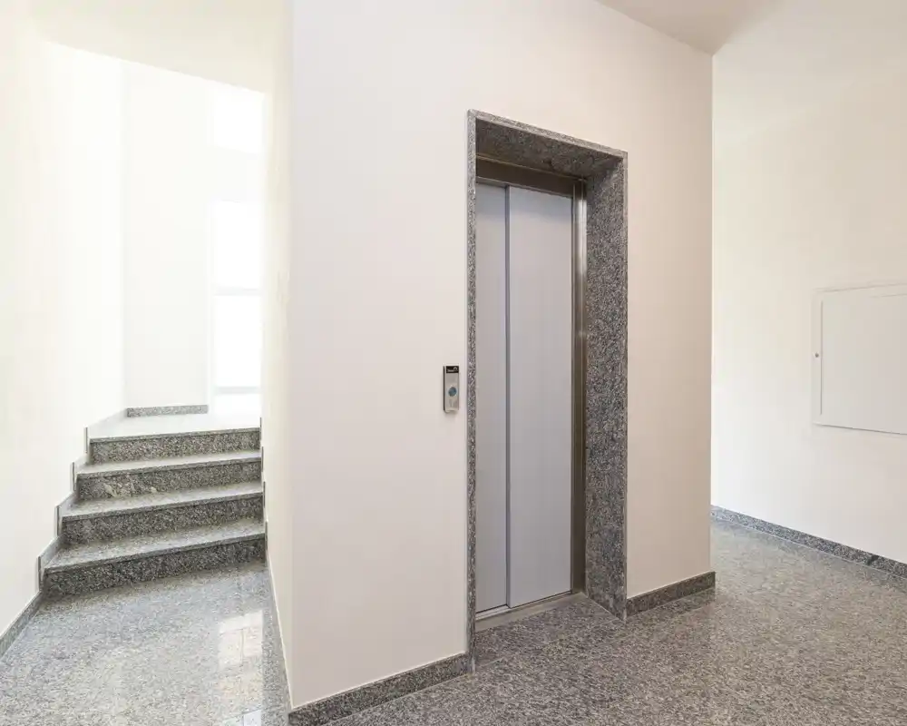 Ingresso/androne luminoso e ben tenuto con ascensore e vano scale adiacente, pavimento in pietra lucidata e pareti bianche pulite.