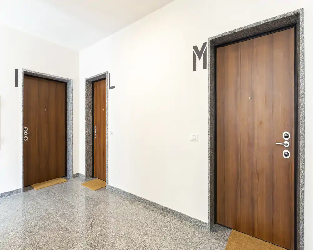 Corridoio/ingresso ben illuminato con tre porte in legno, pavimento in pietra pulito e pareti neutre: area d'ingresso condominiale chiara.