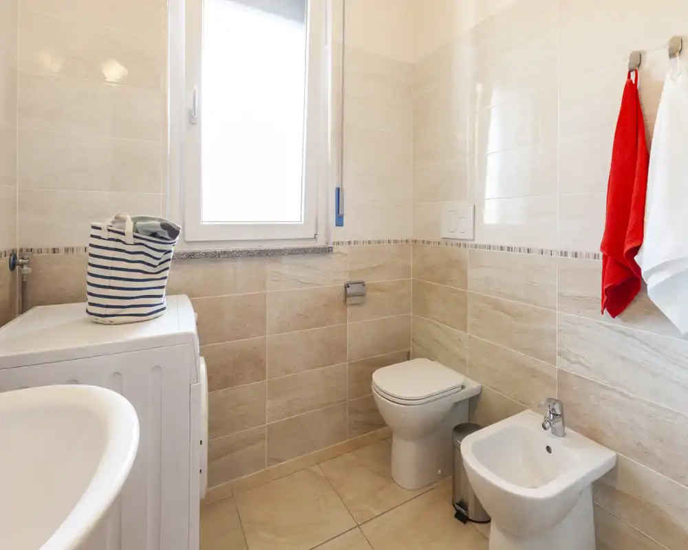 Bagno moderno e luminoso con rivestimenti in ceramica, wc, bidet, lavabo e una lavatrice accanto alla finestra che garantisce luce naturale.