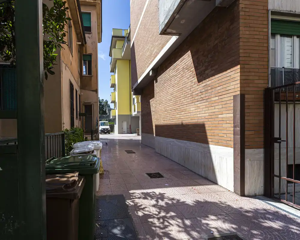 Vicoletto soleggiato tra palazzi residenziali con pavimentazione piastrellata, bidoni della spazzatura e vista verso la strada; luce diurna intensa e cielo blu.