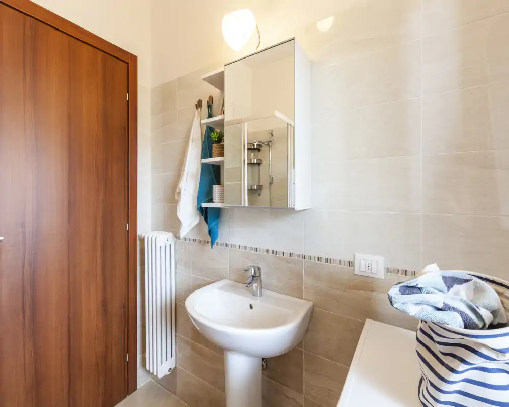 Bagno moderno e luminoso con lavabo a colonna, specchiera contenitore, ganci per asciugamani e piastrelle dai toni neutri.