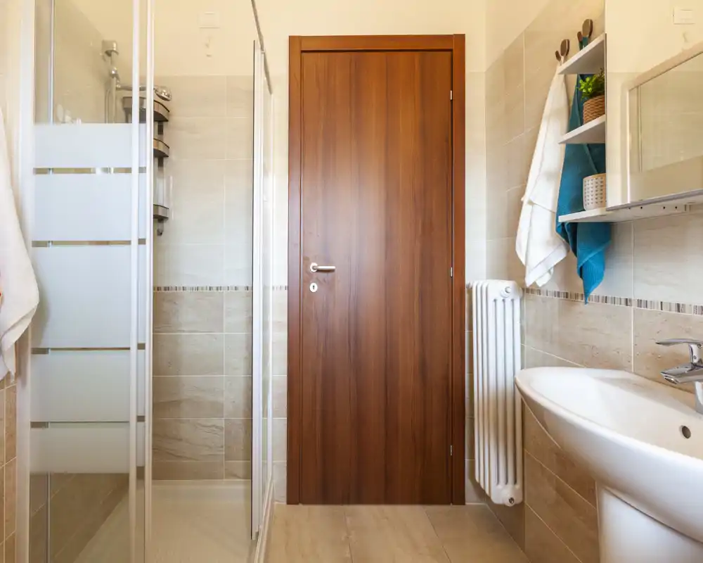 Bagno moderno e pulito con box doccia in vetro, lavabo sospeso, termosifone e piastrelle beige neutre che conferiscono un aspetto caldo e ordinato.