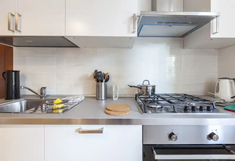Cocina compacta y moderna con encimera de acero inoxidable, vitrocerámica a gas, fregadero y pequeños electrodomésticos; luminosa y ordenada, lista para cocinar.