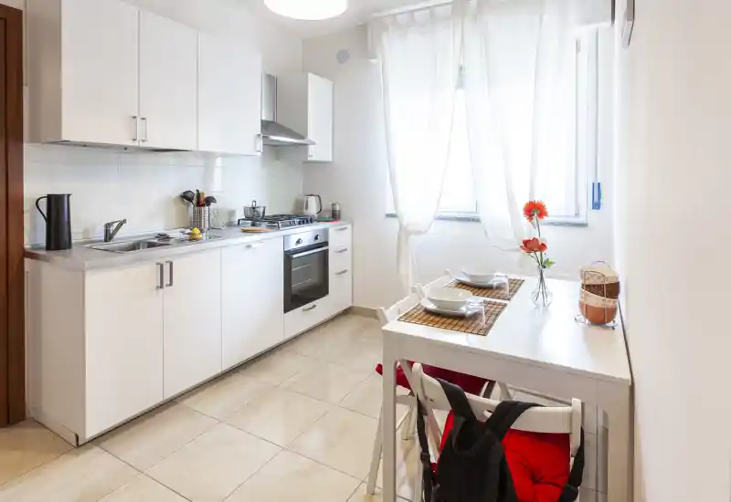 Cocina moderna y luminosa con muebles blancos, horno empotrado y una pequeña mesa para dos; la luz natural y detalles decorativos la hacen acogedora.