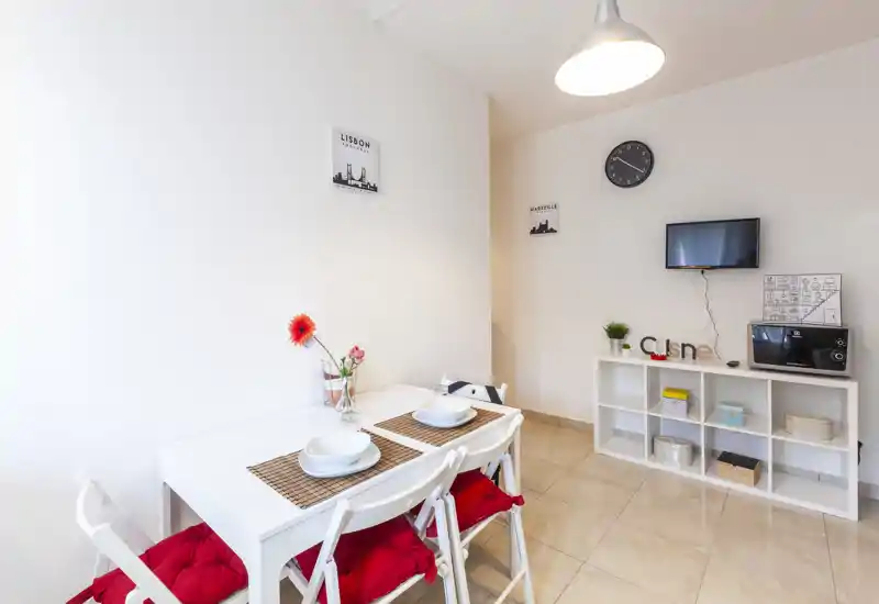 Rincón comedor luminoso y compacto con mesa blanca puesta para dos, cojines rojos y jarrón decorativo — ideal para comidas acogedoras en un piso pequeño.