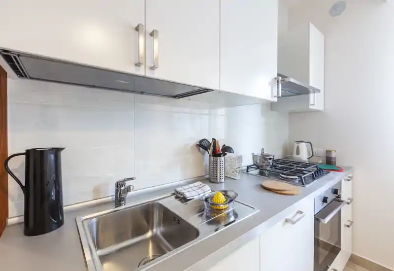 Cocina compacta y moderna con fregadero de acero inoxidable, vitrocerámica de gas y horno empotrado; espacio luminoso y ordenado con pequeños accesorios y muebles blancos contemporáneos.