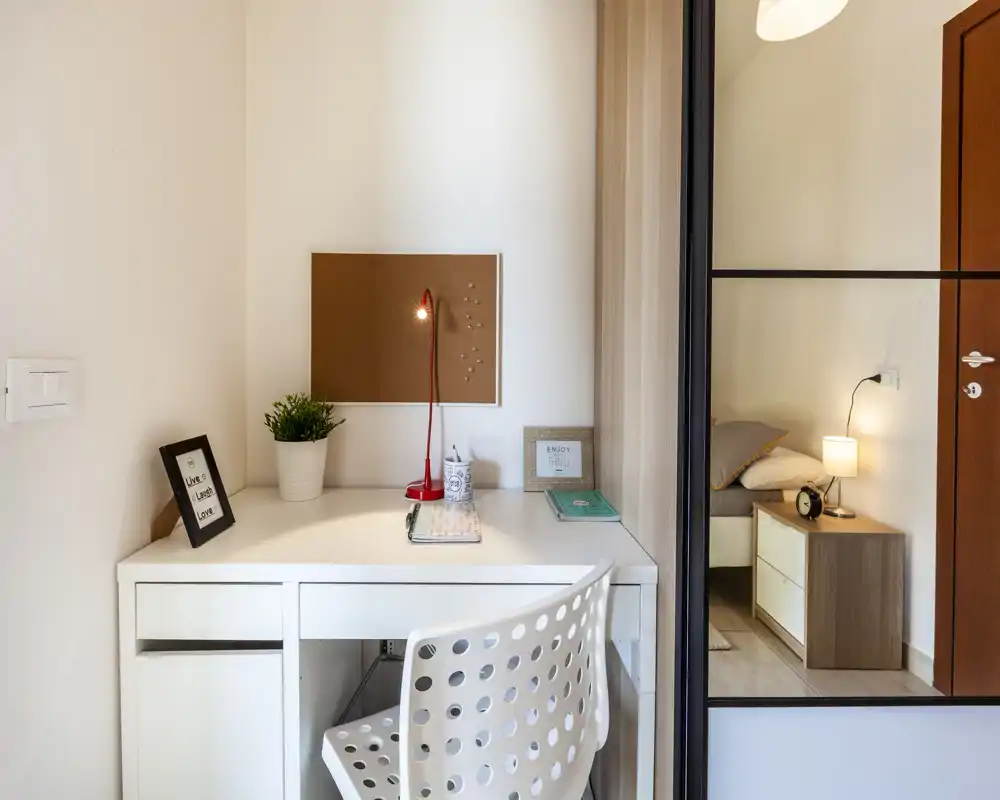 Angolo camera ben illuminato con una scrivania bianca in primo piano e il letto visibile attraverso una porta con specchio. L'ambiente coniuga un’area di lavoro funzionale con dettagli del comodino — lampada, mobiletto e tessili morbidi — dallo stile moderno e pulito.