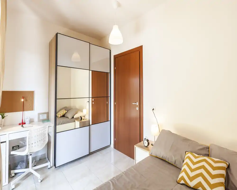 Camera da letto luminosa e moderna con comodo letto matrimoniale, comodino con lampada, guardaroba a specchio e un piccolo scrittoio — perfetta per riposo e studio.