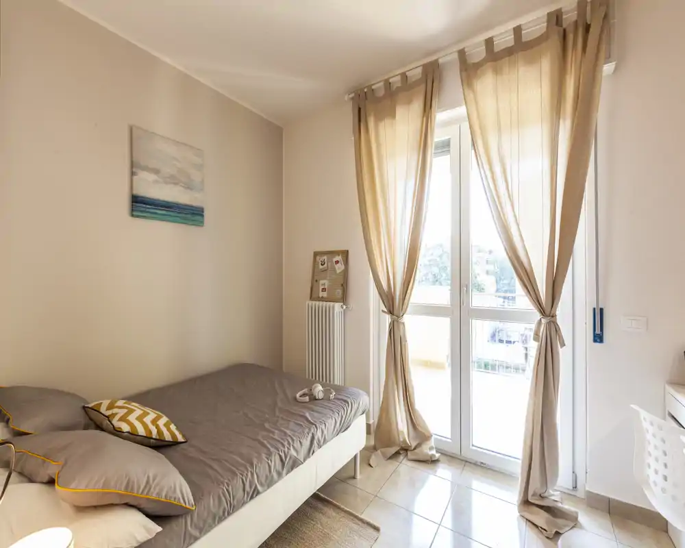 Camera da letto luminosa e accogliente con letto matrimoniale, biancheria grigia, cuscini decorativi e porte-finestre che illuminano la stanza, incorniciate da tende beige.