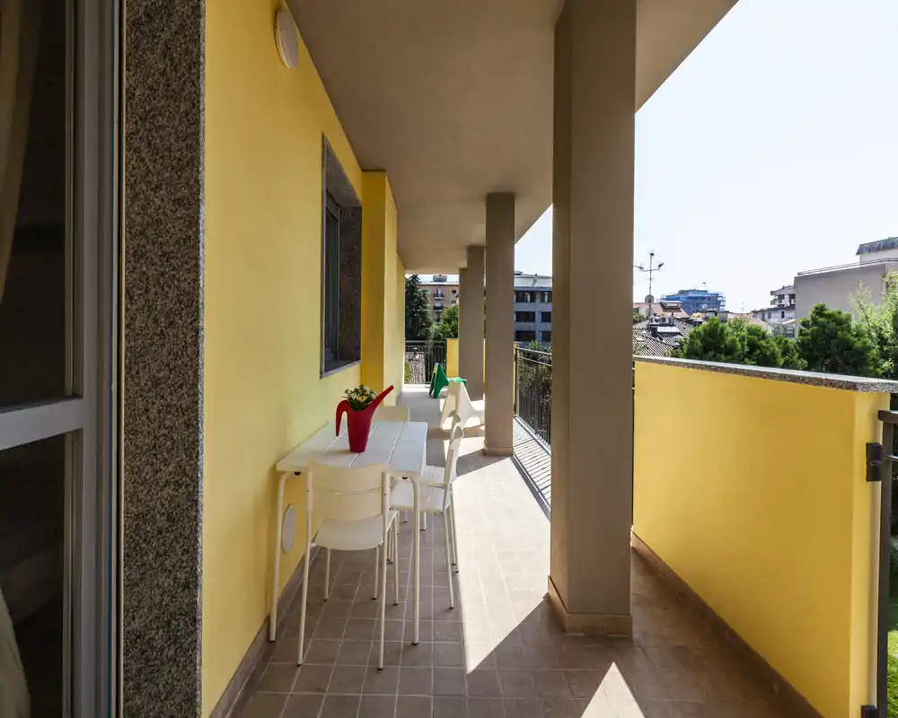 Balcone soleggiato arredato con tavolo lungo, sedie bianche e pianta in vaso; pavimento piastrellato e ringhiera che guarda gli edifici vicini — ideale per pranzi all'aperto e relax.