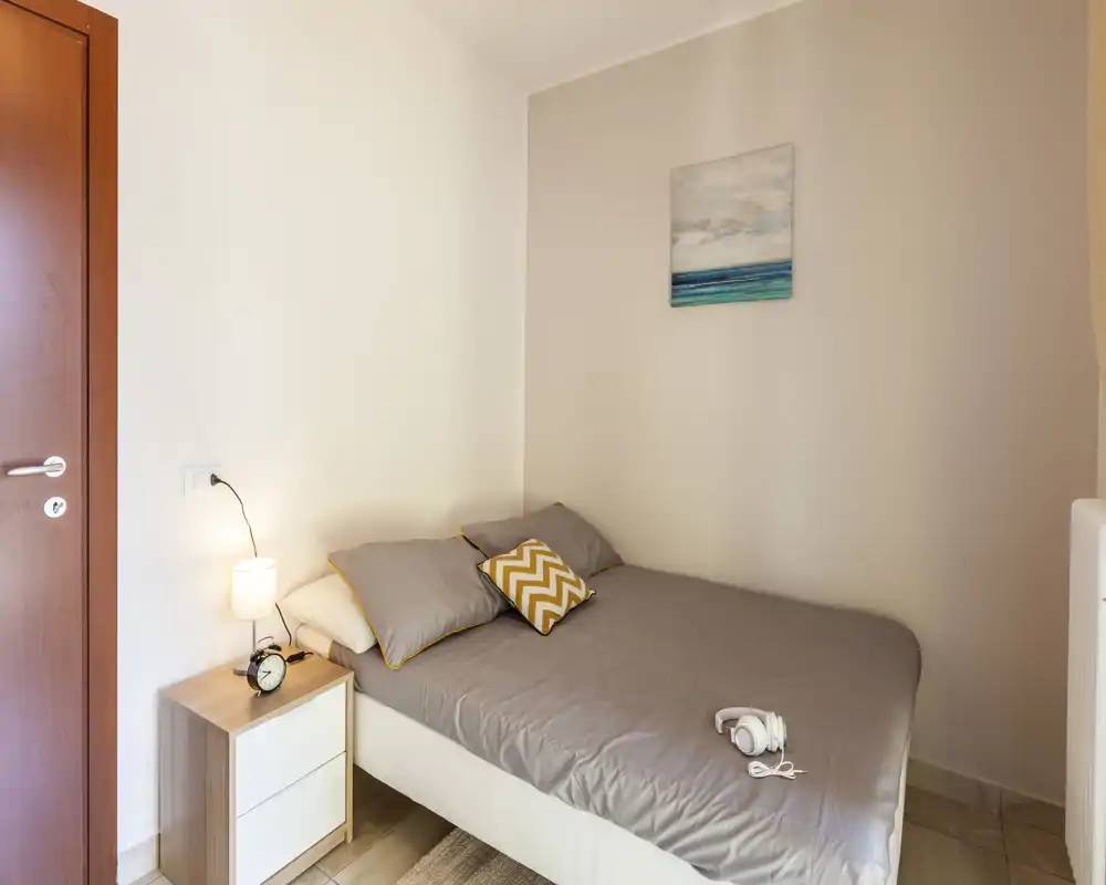 Camera da letto compatta e luminosa con letto matrimoniale, lenzuola neutre, comodino con lampada e cuscino decorativo che crea calore.