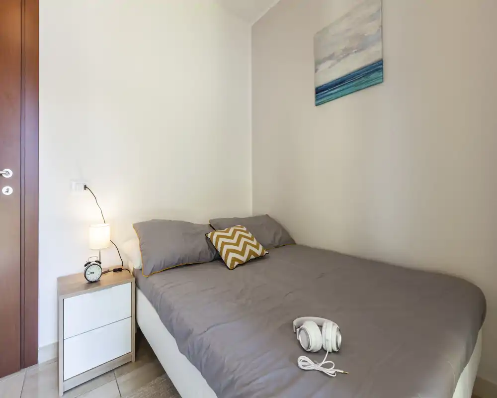 Camera da letto accogliente e ben illuminata con letto matrimoniale con biancheria grigia, cuscino decorativo, comodino con lampada e sveglia — perfetta per un soggiorno rilassante.