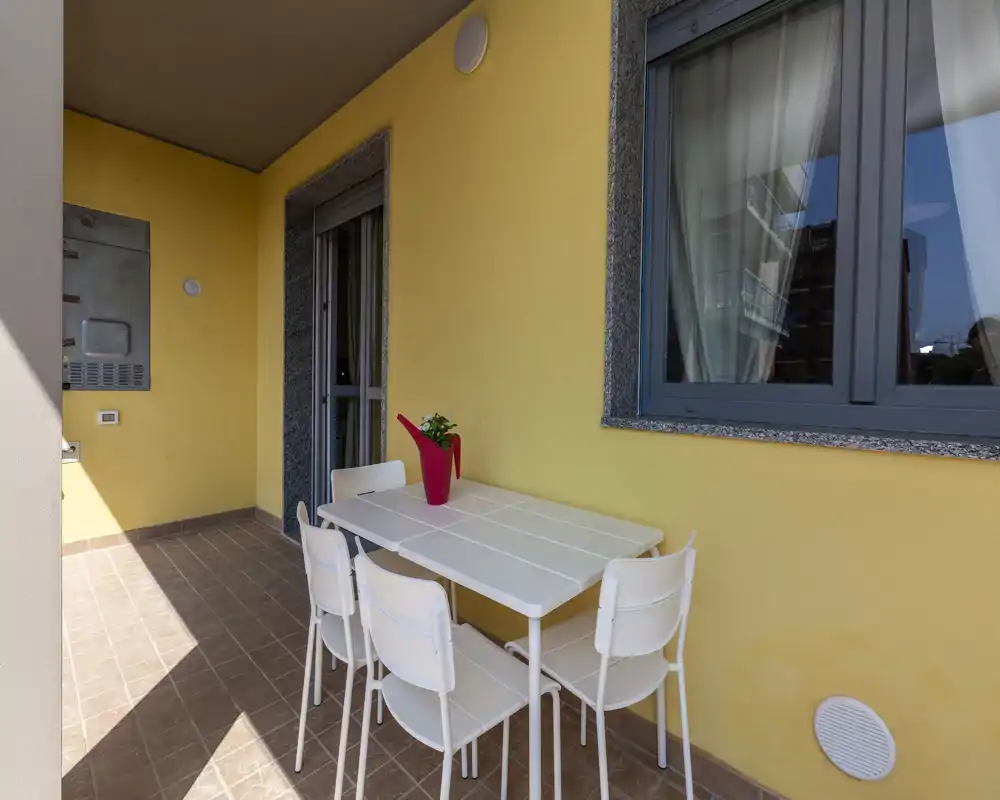 Balcone soleggiato arredato con tavolo bianco e quattro sedie, pareti gialle e piccolo vaso decorativo — ideale per pranzi all'aperto o relax.