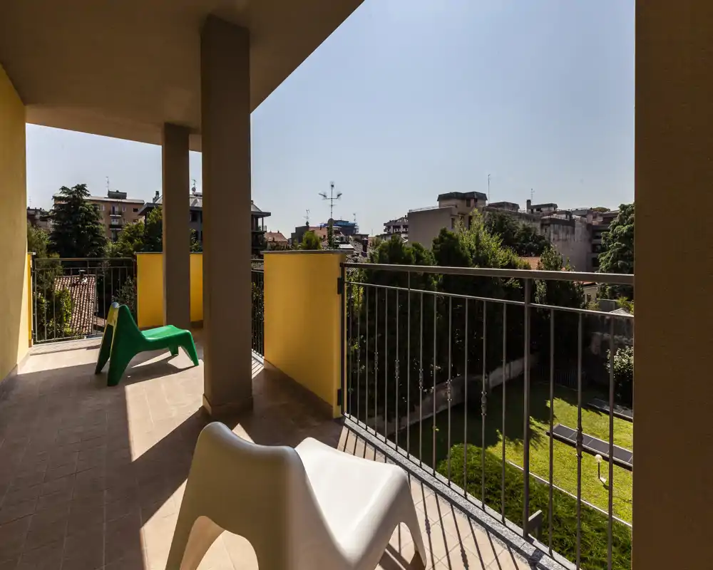 Balcone privato soleggiato con sedute esterne e vista su un cortile alberato e gli edifici circostanti; pavimento piastrellato e ringhiera proteggono lo spazio esterno.