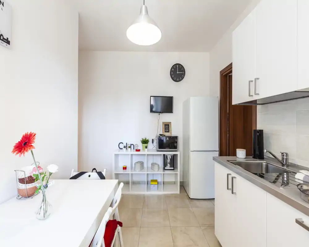 Cucina moderna e luminosa con mobili bianchi, lavello in acciaio, frigorifero e un piccolo tavolo da pranzo con elementi decorativi.