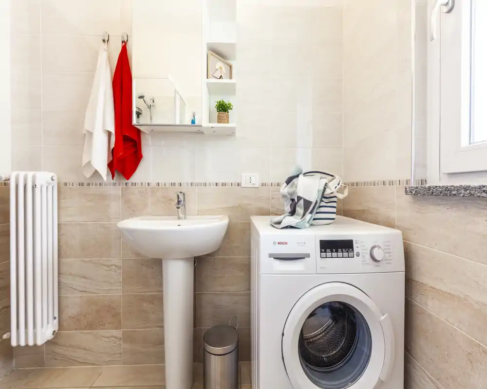 Bagno moderno e luminoso con lavabo a colonna, pensile a specchio, ganci per asciugamani e lavatrice a carica frontale — funzionale e ben illuminato.