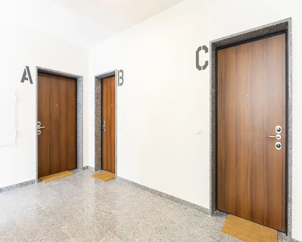 Ingresso condominiale con tre porte numerate, pavimento in pietra pulito e zerbini semplici—corridoio comune luminoso e ben illuminato.