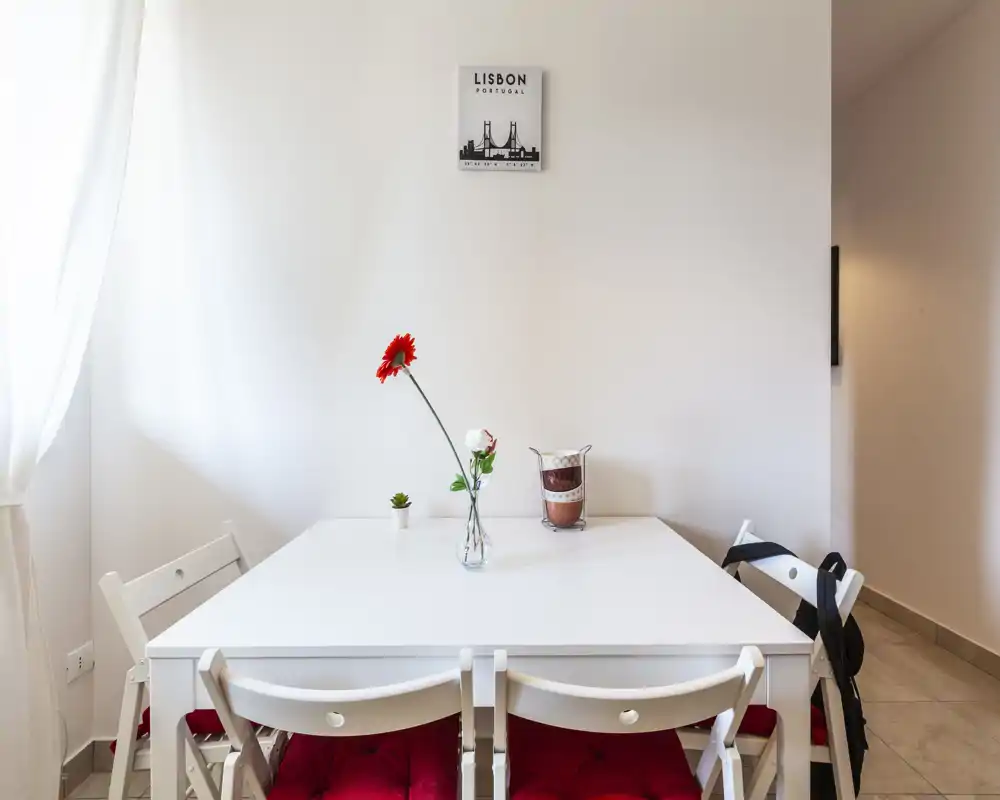 Zona pranzo luminosa e minimal con tavolo bianco, quattro sedie con cuscini rossi e un semplice centrotavola floreale, perfetta per pasti informali.