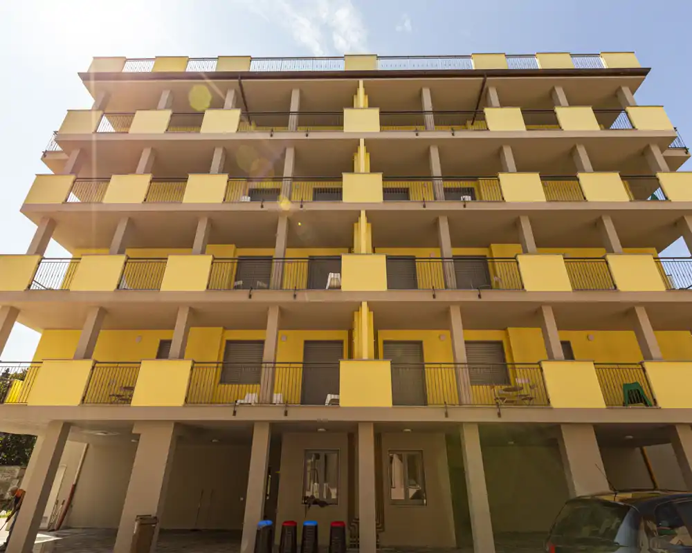 Vista esterna di un moderno edificio residenziale giallo con più balconi aperti e un'area parcheggio coperta al piano terra, fotografato con luce solare intensa.