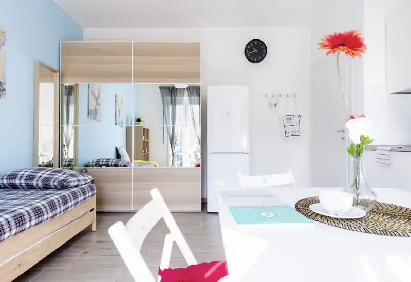 Bright, modern studio-bedroom with a neatly made single bed, mirrored wardrobe and a small dining/work table bathed in natural light.