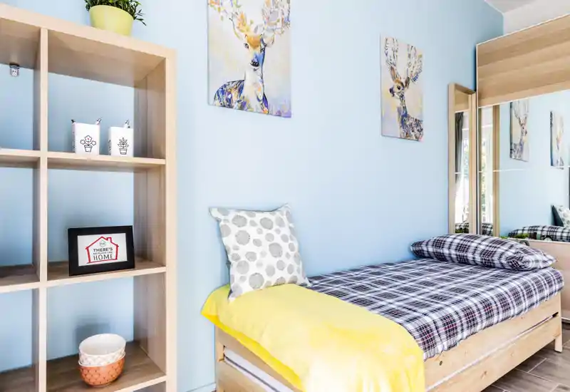 Bright, neatly styled single bedroom with a plaid bed, colorful wall art and a wooden shelving unit — ideal for a cozy rental stay.