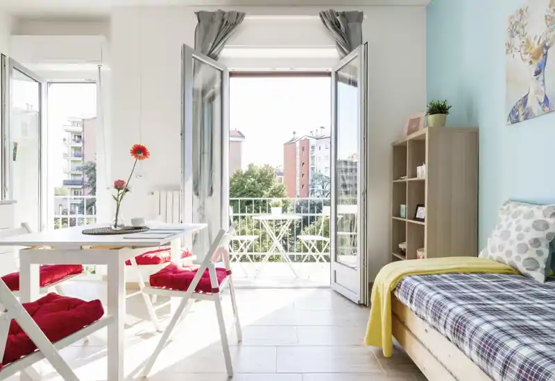 Bright, airy studio corner with an open balcony, a single bed with patterned bedding, and a small dining table with red-cushioned chairs — ideal for a cozy city stay.