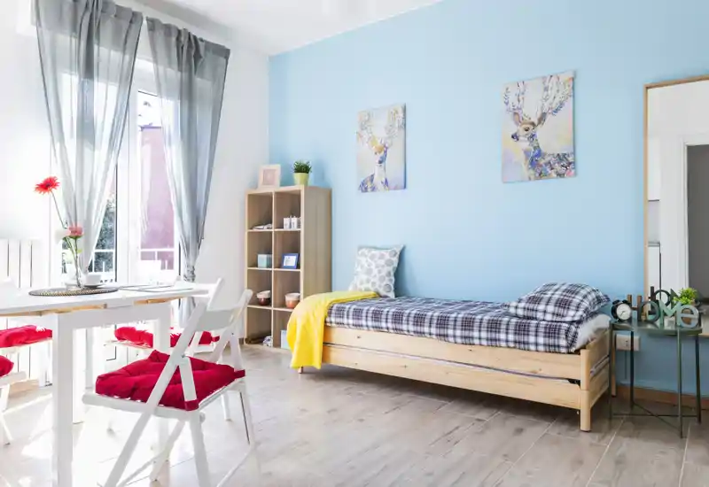 Bright, tidy bedroom with a single wooden bed, cozy textiles and a dining nook by a large sunlit window. Light blue accent wall and decorative prints give the room a fresh, welcoming look — ideal for a compact rental.