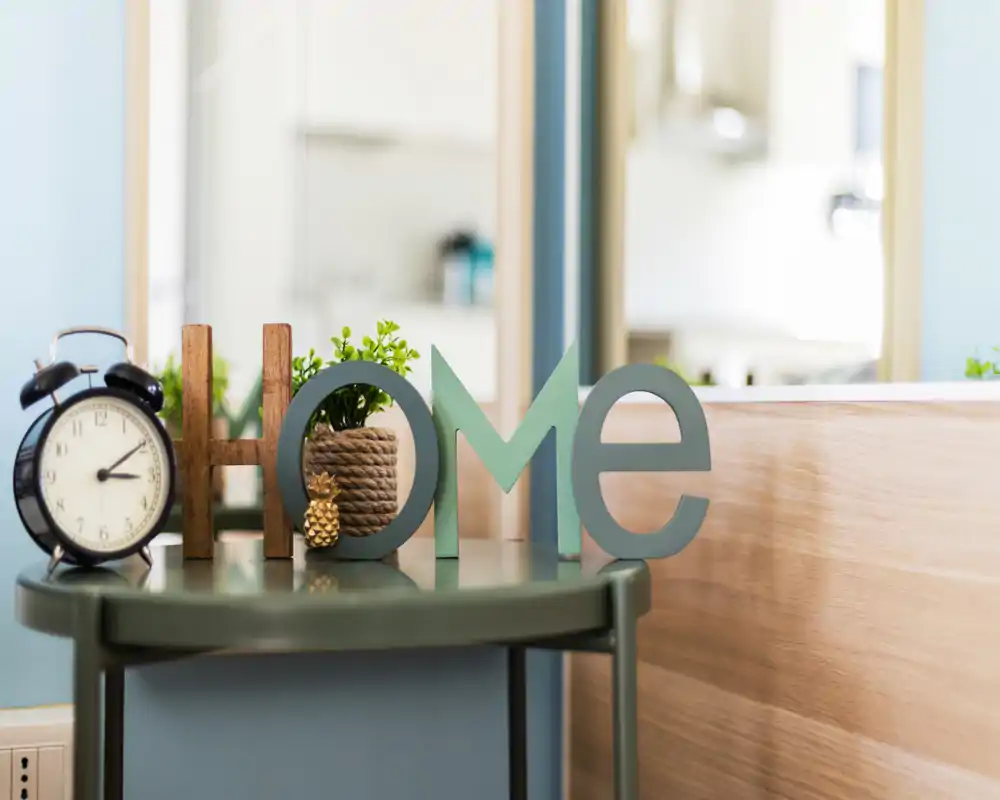 Close-up decorative shot on a bedside table with an alarm clock, small plant and wooden letters spelling “HOME”, set against a light, cozy background.